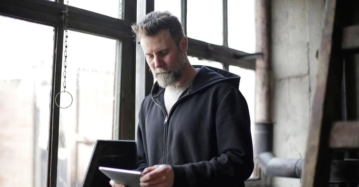Homme barbu en sweat à capuche noir travaillant sur une tablette près de fenêtres industrielles.