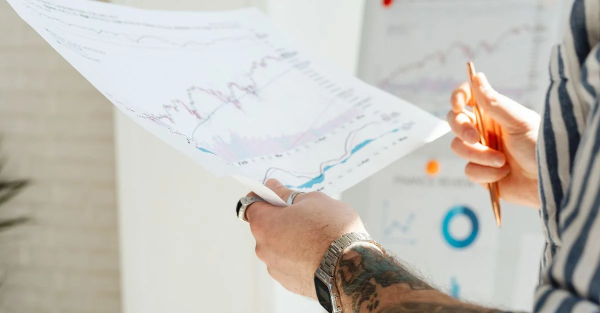 Gros plan sur les mains d'un homme tatoué tenant des graphiques financiers, avec un accent sur l'analyse des données et les tendances du marché boursier.