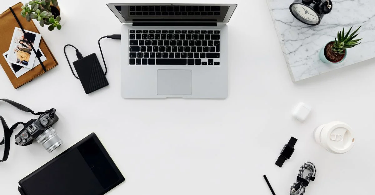 Vue de dessus d'un espace de travail minimaliste avec un ordinateur portable, un appareil photo et divers accessoires électroniques sur une table blanche.