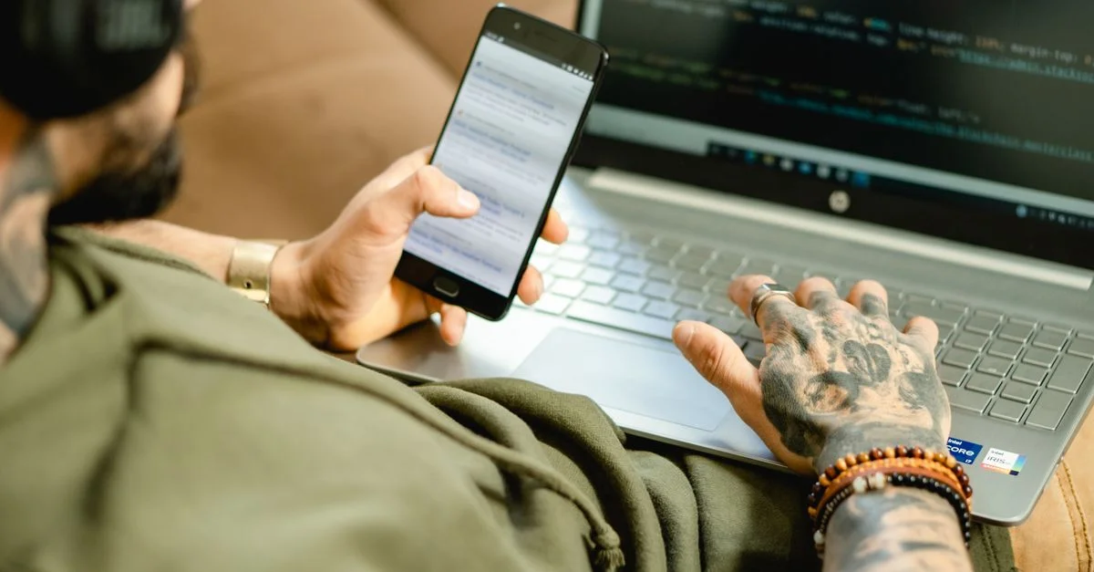 Une personne tatouée engagée dans la programmation sur un ordinateur portable tout en consultant un smartphone.