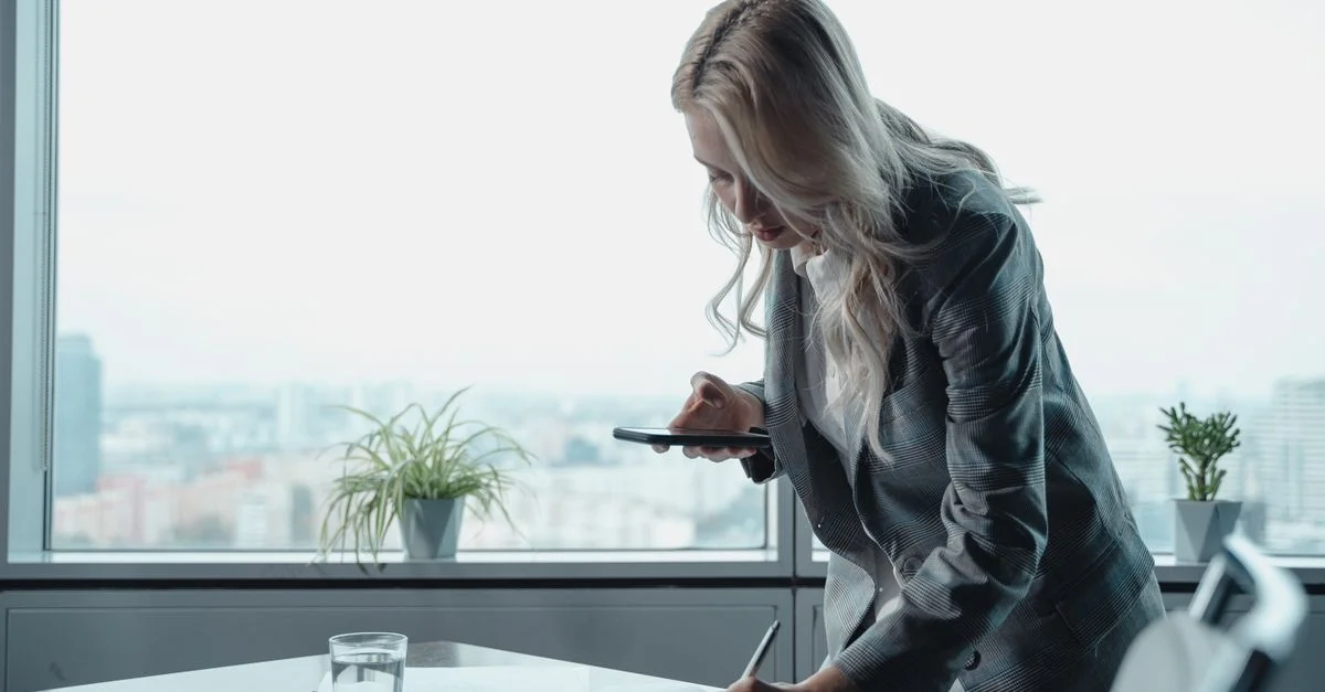 Femme d'affaires avec des cheveux blonds écrivant des notes avec un smartphone dans un bureau moderne.
