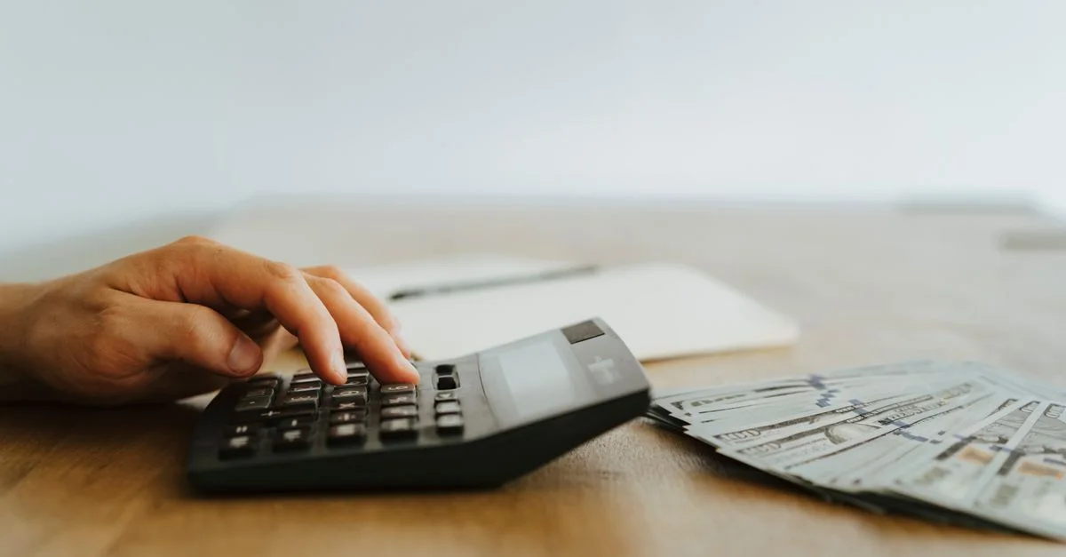 Une main calculant des finances avec une calculatrice à côté de tas de billets en dollars américains.