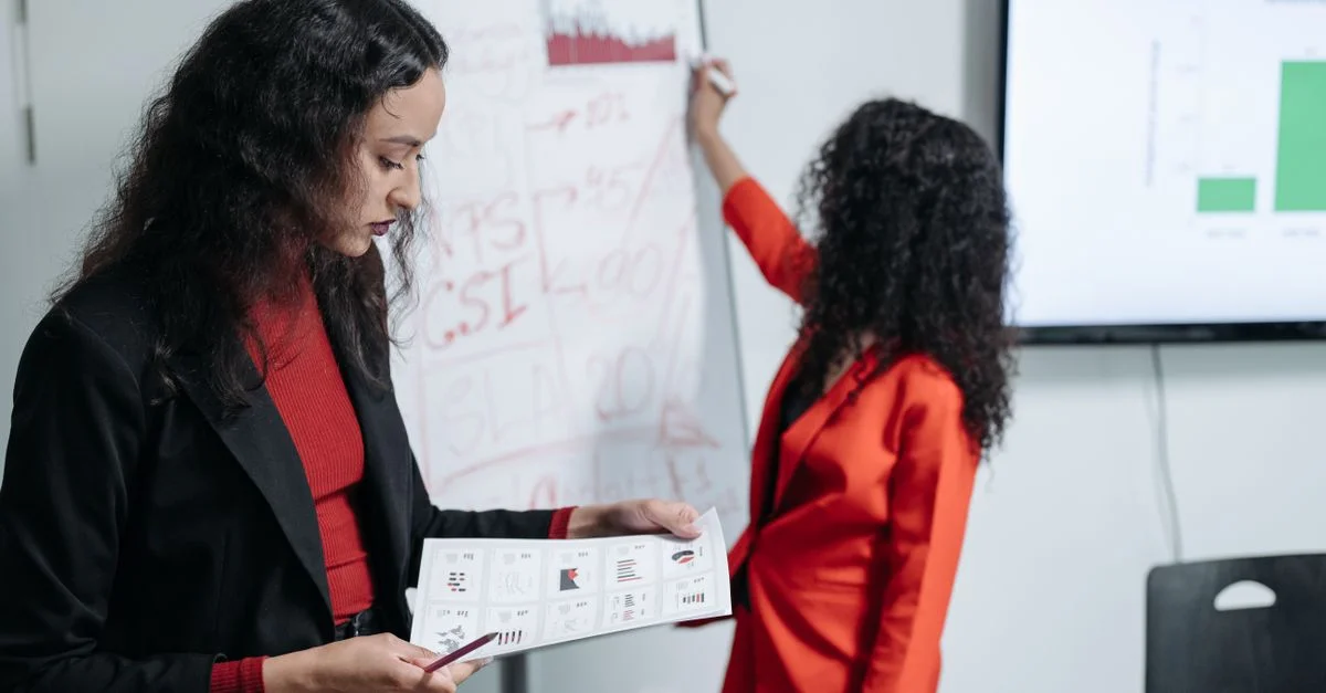 Deux femmes en réunion d'affaires analysant des graphiques et des tableaux de données dans un bureau moderne.